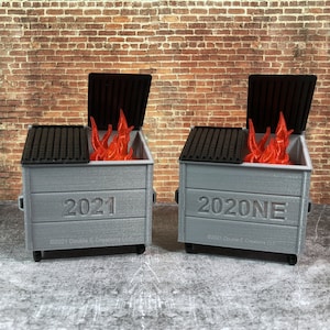 May include: Two miniature grey dumpsters with black lids and red flames inside. The dumpster on the left has the year 2021 printed on the front, and the dumpster on the right has the year 2020NE printed on the front.