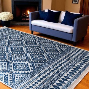 Op de afbeelding: Een rechthoekig vloerkleed met een blauw en wit geometrisch patroon. Het kleed ligt op een houten vloer. Op de achtergrond staat een blauwe bank met marineblauwe kussens, bij een open haard en een raam.