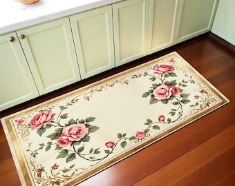 May include: Rectangular cream-colored kitchen rug with a floral rose design. The rug features large pink roses with green leaves and a gold border. The rug is placed on a hardwood floor in front of light green cabinets.
