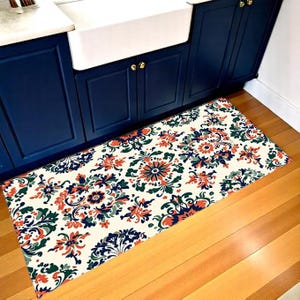 May include: A rectangular kitchen mat featuring a floral design in orange, navy blue, and green on an off-white base. The mat is positioned in front of a navy blue cabinet and a white sink.