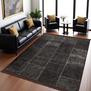 Op de afbeelding: Een zwart lederen patchwork tapijt met een verweerde look. Het tapijt ligt op een houten vloer in een woonkamer met zwarte lederen meubels.