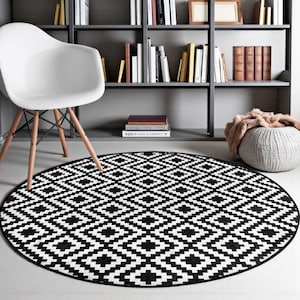 Peut inclure: Tapis rond à motifs géométriques noirs et blancs. Le tapis présente un motif de losanges répétés. Une chaise blanche avec des pieds en bois est à gauche, et une bibliothèque est en arrière-plan.