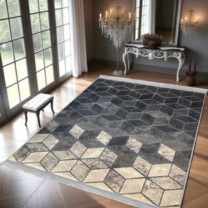 Puede incluir: Alfombra con diseño geométrico y degradado. La alfombra presenta un patrón de cubos repetidos en tonos grises, negros y beige. Tiene un borde con flecos y está colocada sobre un suelo de madera.