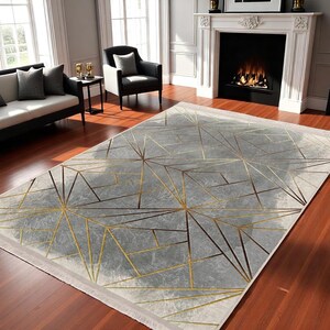 May include: A gray and gold geometric area rug with a modern design. The rug has a white border and is made of a soft, plush material.