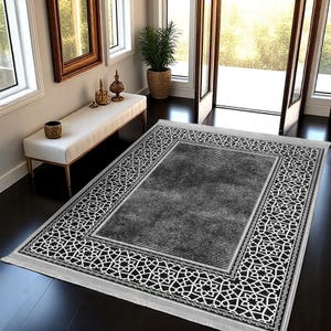 Puede incluir: Una alfombra rectangular con un centro gris y un borde geométrico en blanco y negro. La alfombra tiene flecos en los extremos cortos y está colocada sobre un suelo de madera oscura. La alfombra está en una habitación bien iluminada con un banco y una planta en maceta.
