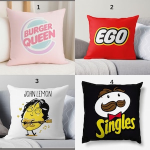 May include: Four decorative cushions with pop culture designs. One pink cushion features "BURGER QUEEN" text. A red cushion displays "EGO" in a white and yellow logo. A white cushion has a cartoon "JOHN LEMON" playing guitar. A black cushion shows a cartoon character with "Singles" text.