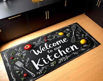 May include: Black rectangular kitchen mat with the words "Welcome to our Kitchen" in white script. The mat features illustrations of a spatula, whisk, lemons, and berries. The mat is on a wooden floor.