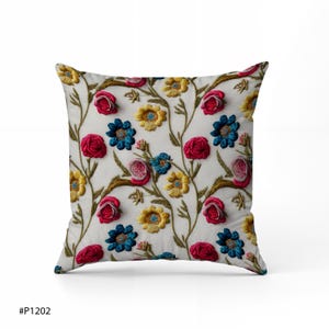 May include: Square decorative pillow with an embroidered floral pattern. The design features red, blue, and yellow flowers with green stems on a white background. The pillow is a square shape and appears to be made of a soft fabric.