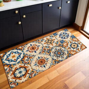 Op de afbeelding: Een rechthoekige vloermat met een herhalend tegelpatroon in crème, blauw en oranje tinten. De mat ligt voor zwarte kasten met gouden handgrepen, op een houten vloer. Het ontwerp bevat bloemen en geometrische motieven.