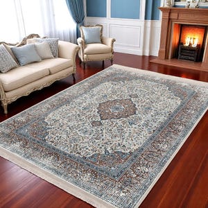 May include: A cream-colored rug with a brown and blue floral pattern. The rug has a fringe border and is laid out on a hardwood floor.