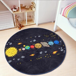 May include: A round, blue and black rug with a solar system design. The rug features a yellow sun with a smiling face, planets with smiling faces, and stars. The text on the rug includes "Sun", "Mercury", "Venus", "Earth", "Mars", "Jupiter", "Saturn", "Uranus", "Neptune", "Comet", and "Constellation".