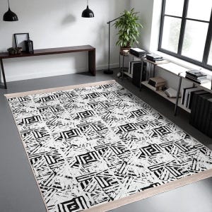 Può includere: Un tappeto rettangolare con un motivo geometrico in bianco e nero. Il tappeto presenta un design ripetuto di forme angolari e linee su sfondo bianco. Ha un bordo beige ed è posizionato su un pavimento grigio. Arredamento per la casa.