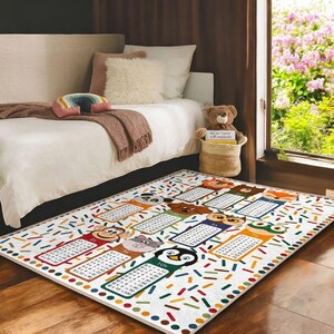 Könnte beinhalten: Ein weißer und bunter Teppich mit einem darauf gedruckten Einmaleins. Der Teppich zeigt Comic-Tiere und bunte Konfetti. Der Teppich liegt auf einem Holzboden in einem Raum mit einem Bett und einem Teddybär.