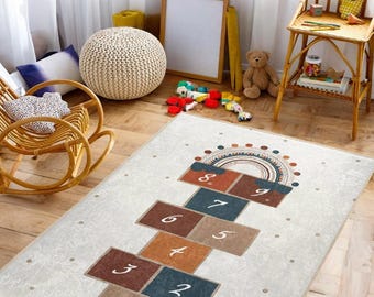 Puede incluir: Una alfombra de juego blanca y beige con un diseño de rayuela con los números del 1 al 9. Los cuadrados de la rayuela son marrones, azules y naranjas. Un arcoíris con un diseño de mandala está en la parte superior de la alfombra.