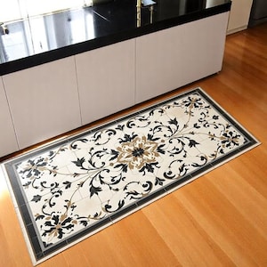 May include: Rectangular floor mat with a floral design. The mat features a cream-colored background with black, brown, and gray floral patterns. A black and gray border frames the design. The mat is placed on a wooden floor.