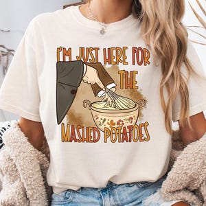 Può includere: T-shirt color crema con la scritta "I'M JUST HERE FOR THE MASHED POTATOES". La grafica mostra una mano che sbatte il purè di patate in una ciotola. Il design ha accenti marroni e arancioni.