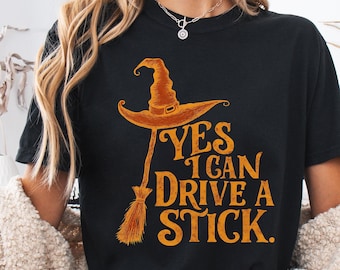 Puede incluir: Camiseta negra con un diseño de Halloween. El diseño presenta un sombrero de bruja y una escoba con el texto "YES I CAN DRIVE A STICK" en naranja. La camiseta la lleva una persona.