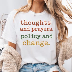 以下が含まれることがあります： 「thoughts and prayers. policy and change.」と赤、緑、金色の文字で書かれた白いTシャツ。長い金髪と青いジーンズのショートパンツを履いた人が着ています。