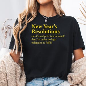 May include: Black t-shirt with the text "New Year's Resolutions" in yellow. The definition below reads "(n.) Casual promises to myself that I'm under no legal obligation to fulfill." The shirt is worn by a person.