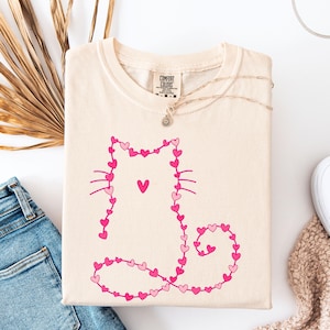 May include: Cream-colored t-shirt featuring a pink heart-shaped cat design. The cat's body is outlined with small hearts, with a larger heart on its chest. The shirt is folded, with a necklace and white shoes visible.