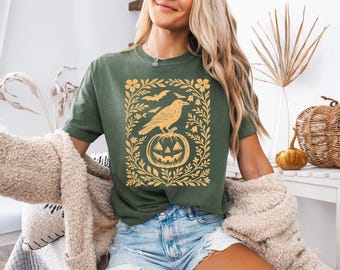 Puede incluir: Camiseta verde con una ilustración dorada de un cuervo sobre una calabaza, rodeada de motivos florales y murciélagos. El diseño está dentro de un marco rectangular. Parte de una colección temática de Halloween.