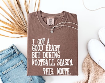 Op de afbeelding: Een bruin t-shirt met de tekst "I GOT A GOOD HEART BUT DURING FOOTBALL SEASON. THIS. MOUTH." wordt weergegeven op een witte ondergrond. Een zilveren ketting, een blauwe jeans en witte sneakers zijn ook zichtbaar.