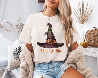 Op de afbeelding: Crèmekleurig T-shirt met een afbeelding van een heksenhoed, een spin en de tekst "IF THE HAT FITS" in bruin. De hoed is zwart, groen en paars met een bruine riem. Een casual, Halloween-thema T-shirt.