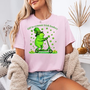 May include: Pink t-shirt with a graphic of a green frog wearing a leprechaun hat and riding a green scooter. The text "Off to commit Irish foolery" is above the frog. Gold coins and shamrocks are also in the design.