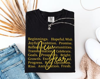 May include: Black t-shirt with gold text listing positive words like "Beginnings," "Ambition," and "Growth." A silver necklace with a circular pendant rests on the shirt. The shirt is paired with blue jeans and white shoes.