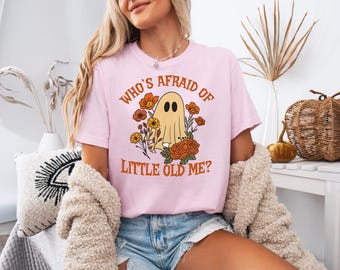 Könnte beinhalten: Hellrosa T-Shirt mit dem Text "WHO'S AFRAID OF LITTLE OLD ME?" und einem Cartoon-Geist, der von Blumen umgeben ist. Der Geist hat zwei schwarze Augen und trägt einen Blumenkranz. Das T-Shirt hat eine hellrosa Farbe.
