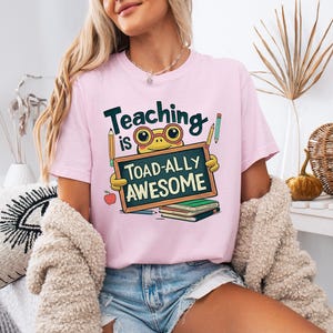 May include: Pink t-shirt with the phrase "Teaching is Toad-ally Awesome" in black and green. The graphic features a cartoon frog wearing glasses holding a chalkboard. Pencils and an apple are also included in the design.