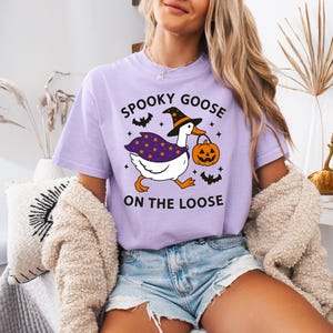 May include: A light purple t-shirt with the text "SPOOKY GOOSE ON THE LOOSE". The graphic features a white goose wearing a witch hat and cape, carrying a jack-o'-lantern. Black bats and stars surround the goose, perfect for Halloween.