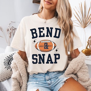 Op de afbeelding: Crèmekleurig T-shirt met de woorden "BEND & SNAP" in marineblauwe blokletters boven en onder een voetbalafbeelding. De voetbal is oranje en bruin met een marineblauw ampersand. Het shirt wordt gedragen met een spijkerbroekshort.