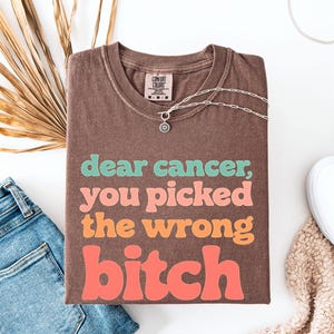 Puede incluir: Una camiseta marrón con el texto "dear cancer, you picked the wrong bitch" en verde azulado, rosa, naranja y coral. Un collar plateado, vaqueros azules y zapatillas blancas también están en la imagen.