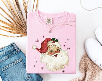 May include: A light pink t-shirt with a vintage Santa Claus design. The graphic shows Santa with a red hat, white beard, and a smiling face. The shirt is folded, with blue jeans, white shoes, and a necklace nearby.