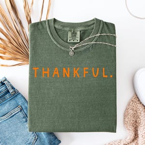 May include: An olive green t-shirt with the word "THANKFUL" in orange lettering. The shirt is paired with blue jeans, white slip-on shoes, and a silver necklace. A dried palm leaf adds a natural touch.