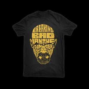 May include: Black t-shirt with a yellow graphic design. The design features the words "Breaking Bad" and "I am the danger" arranged to form a stylized face. The text is in a distressed, textured font.