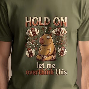 Può includere: T-shirt verde oliva con un capibara dei cartoni animati che medita e la scritta "HOLD ON let me overthink this". Il design include punti interrogativi e fumetti di pensiero, creando una grafica umoristica e riconoscibile.