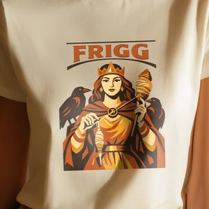 Può includere: T-shirt color crema con la scritta "FRIGG" sopra una grafica di una donna incoronata che tiene un fuso, affiancata da due uccelli neri. La donna indossa un mantello e un ciondolo d'oro. Il design è nei toni del marrone, arancione e oro.