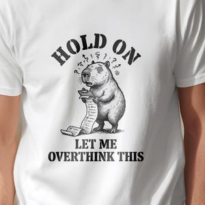 Può includere: T-shirt bianca con un'illustrazione in bianco e nero di un capibara che tiene una lunga lista. Il testo "HOLD ON" è sopra e "LET ME OVERTHINK THIS" sotto. Il capibara ha un'espressione preoccupata.