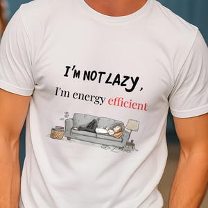 Può includere: T-shirt bianco con la scritta "I'M NOT LAZY, I'm energy efficient" in nero e rosso. La maglietta presenta un'illustrazione a cartoni animati di una persona che si rilassa su un divano. La persona indossa una maglietta bianca e pantaloni scuri.