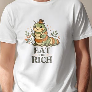 Può includere: T-shirt bianca con un bruco dei cartoni animati con un cappello a cilindro e una forchetta e un coltello. Il testo "EAT THE RICH" è stampato sotto. Il bruco è verde, arancione e marrone. Un capo d'abbigliamento casual.