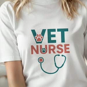 Può includere: T-shirt bianca con le parole "VET NURSE" in verde acqua e rosso, con impronte di zampe e una grafica di stetoscopio. La maglietta è a girocollo.