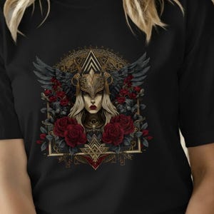 Può includere: T-shirt nero con una grafica dettagliata di una donna con un elmo dorato alato, circondata da rose rosse. Il design ha uno stile vintage e artistico, con dettagli intricati e un'estetica gotica.