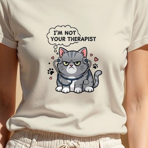 Può includere: Una maglietta beige chiaro con un gatto dei cartoni animati con uno stetoscopio e la scritta "I'M NOT YOUR THERAPIST" in un fumetto. Il gatto è grigio con un'espressione arrabbiata. Sono visibili anche piccole impronte di zampe e cuori.