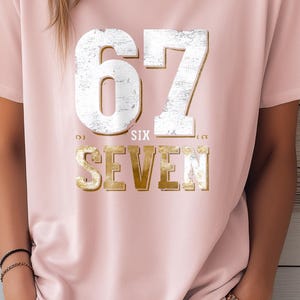 Può includere: T-shirt rosa chiaro con il numero "67" in un carattere bianco e oro invecchiato. La parola "SIX" è sopra "SEVEN" in oro. La maglietta è in stile casual.