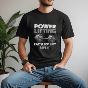 Pode incluir: Camiseta preta com o texto "POWER LIFTING" acima de um gráfico de barra e as palavras "EAT SLEEP LIFT REPEAT" em branco. A camiseta é usada por uma pessoa sentada.