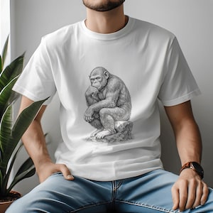 Può includere: T-shirt bianco con un'illustrazione in bianco e nero di uno scimpanzé pensieroso seduto su una roccia. La maglietta è realizzata in un materiale morbido e confortevole.