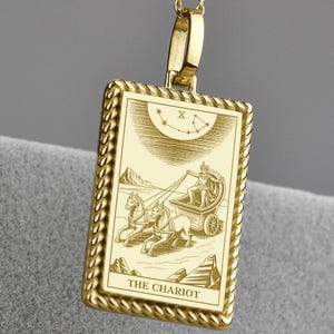 Könnte beinhalten: Goldfarbene Anhängerkette mit einem rechteckigen Anhänger mit einer detaillierten Gravur einer Wagenszene. Der Anhänger hängt an einer filigranen Kette mit kleinen, runden Perlen. Die Worte "THE CHARIOT" stehen unten auf dem Anhänger.