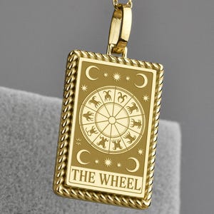 Könnte beinhalten: Goldfarbene Halskette mit einem rechteckigen Anhänger, auf dem die Worte "THE WHEEL" stehen. Der Anhänger zeigt ein detailliertes Design eines Rades mit Tierkreiszeichen und Monden. Die Kette ist filigran.